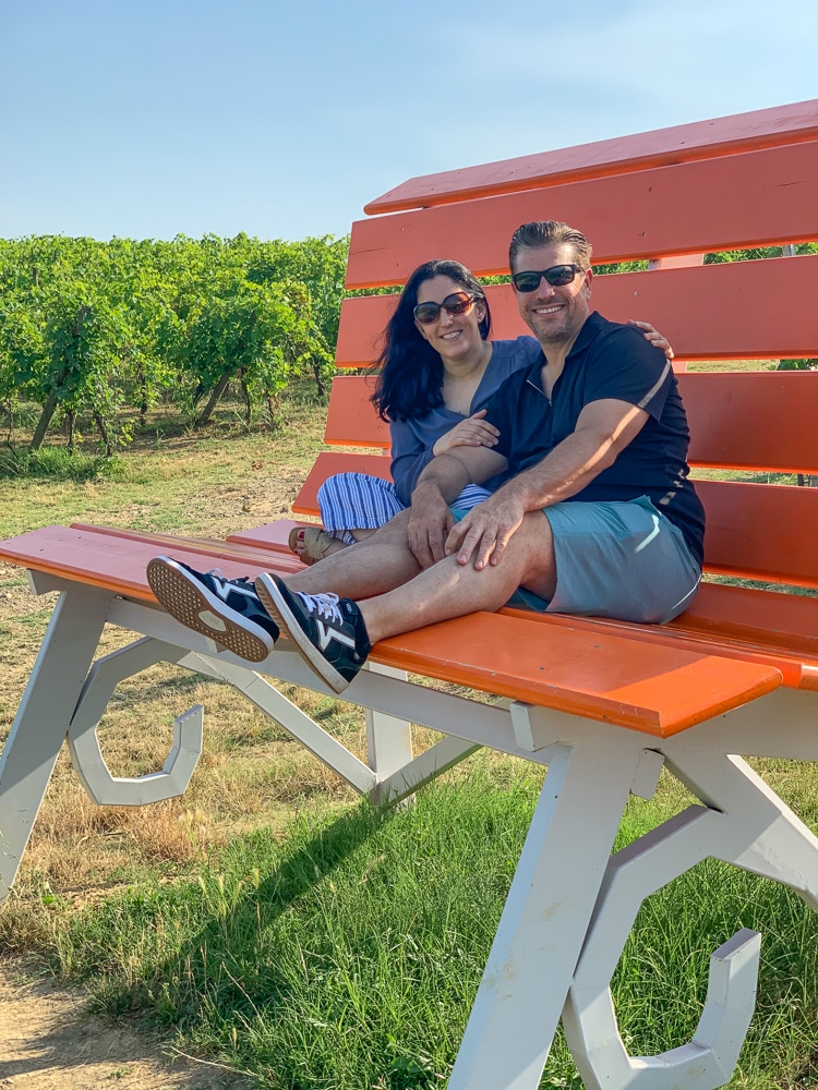2 people sitting on a big orange bench with vineyard vines in the background